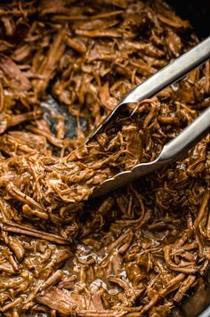 two tongs are being used to stir shredded meat in a pan with brown sauce