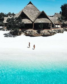 two people are walking on the beach in front of some huts with thatched roofs