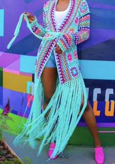 a woman posing in front of a colorful wall wearing a blue fringe jacket and pink shoes