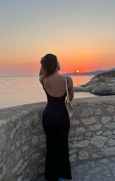 a woman in a black dress looking out at the sunset over the ocean with her back to the camera