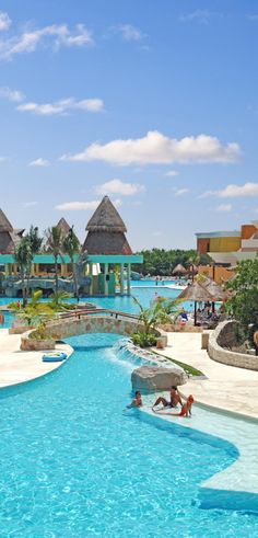 people are swimming in an outdoor pool with thatched huts on the beach behind them