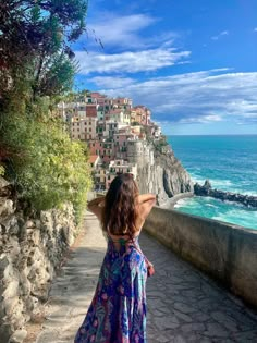 a woman standing on the side of a road looking out at the ocean and buildings