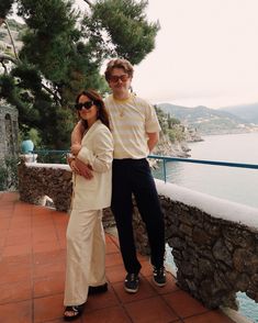 a man standing next to a woman on top of a stone wall near the ocean