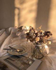 a glass vase filled with dried flowers on top of a white sheet covered bed next to an open book