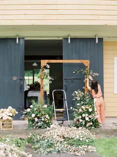 a woman standing in front of a house with flowers on the ground and an open door