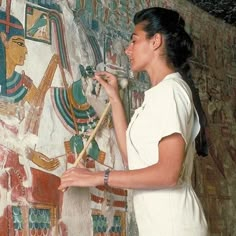 a woman in white shirt painting on wall with egyptian paintings behind her and another person holding a paintbrush