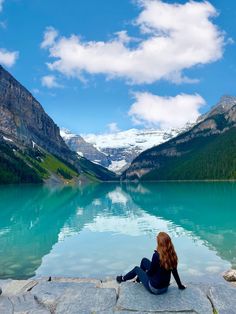 a woman sitting on the edge of a lake with mountains in the background and clouds in the sky