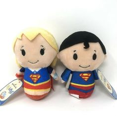 two small stuffed toys are sitting next to each other on a white surface, one has a tag in its mouth and the other is wearing a superman costume