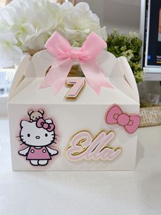 a hello kitty themed gift box with pink bows and gold lettering on the front, sitting on a table next to white flowers