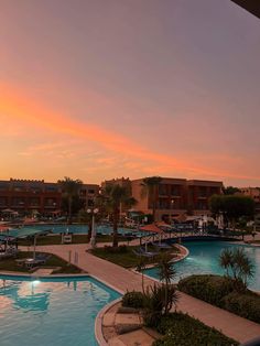 an outdoor swimming pool surrounded by palm trees and other buildings at sunset or dawn with the sun setting in the distance