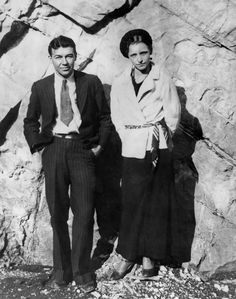 an old black and white photo of two people standing next to each other in front of a rock wall