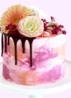 a pink and white cake with chocolate drips, flowers and greenery on top