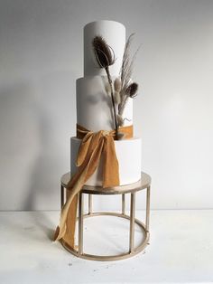 a three tiered white cake with gold ribbon and feathers on top, sitting on a stand