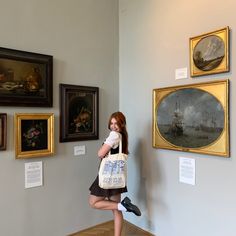 a woman is posing in front of some paintings and holding a tote bag with her right leg