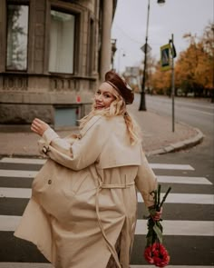 a woman is crossing the street with flowers in her hand and wearing a trench coat