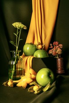 an arrangement of fruit and flowers on a black background with a yellow curtain behind it