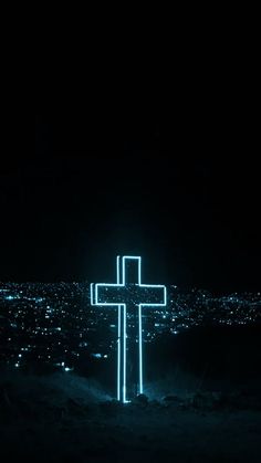 a cross lit up in the dark with city lights behind it on a hill at night