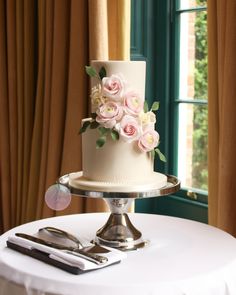 a white cake with pink flowers on top sitting on a table next to a window