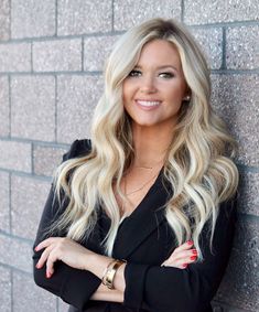 a woman leaning against a brick wall with her arms crossed and smiling at the camera