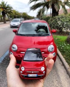 a hand holding a toy car in front of a red car on the side of the road