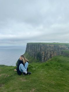 a woman sitting on top of a lush green hillside next to the ocean with cliffs in the background