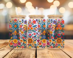 three tumblers with colorful flowers on them sitting on top of a wooden table in front of a blurry background