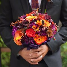 a man in a suit and tie holding a bouquet with oranges, purples and red flowers