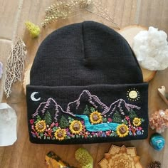 a black beanie hat with sunflowers and mountains on it next to other items