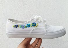 someone is holding their white tennis shoe with blue flowers on the side and green leaves on the bottom