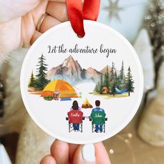 someone holding up a christmas ornament that says, let the adventure begin with two people sitting at a campfire