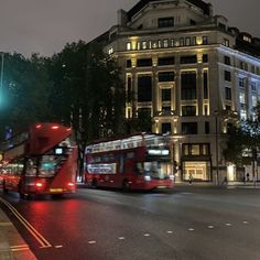 two red double decker buses driving down the street at night time, with buildings in the background