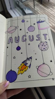 a hand holding an open notebook with space drawings on the pages and words august above it