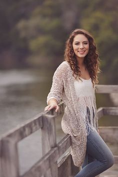 a beautiful young woman leaning on a wooden fence next to the water with her hands in her pockets