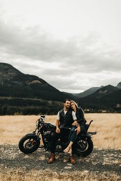 a man and woman sitting on a motorcycle in the middle of a field with mountains behind them