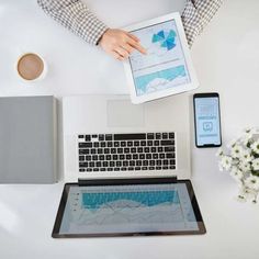 a person holding an ipad next to a laptop on a desk with flowers and other items