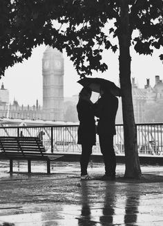 two people standing under an umbrella in the rain next to a bench and clock tower