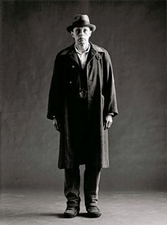 black and white photograph of man in trench coat standing with hat on his head, looking at the camera