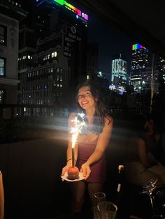 a woman holding a birthday cake with lit candles on it in front of a cityscape