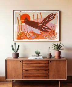 a painting hanging on the wall above a wooden cabinet with two potted plants in front of it