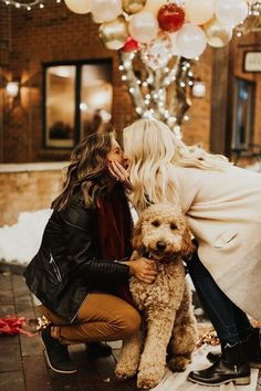 two women kissing and hugging their dog in front of christmas lights on the street with balloons behind them