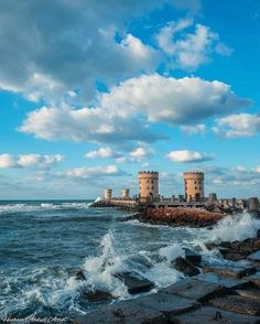 waves crashing against the rocks near an ocean shore with two buildings in the background and blue sky