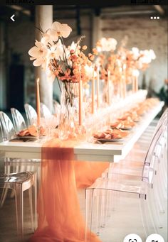 a long table with clear chairs and flowers in vases on the top is surrounded by candles