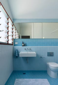 a blue tiled bathroom with a toilet and sink under a window that is open to let in light