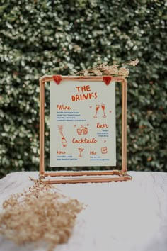 a sign that is sitting on top of a white table cloth with some plants in the background