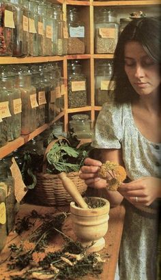 a woman is looking at some herbs in a jar on a table next to a shelf with jars