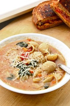 a white bowl filled with pasta soup next to a sandwich on a cutting board and knife