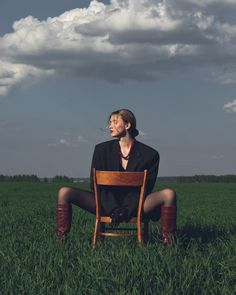 a woman sitting on top of a wooden chair in a green field under a cloudy sky