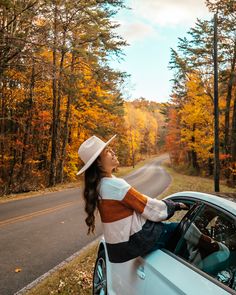 a woman sitting on the hood of a car in front of trees with fall foliage