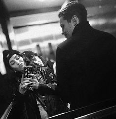 a man and woman standing next to an escalator looking at a cell phone