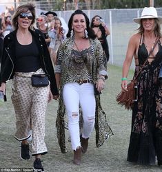 three women are walking together in the grass at an outdoor music festival, one is wearing a black top and white pants